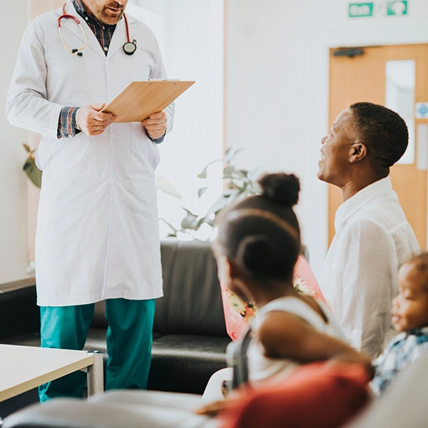 A doctor in a white coat holds a clipboard while talking to a group of attentive patients in a cozy waiting area.