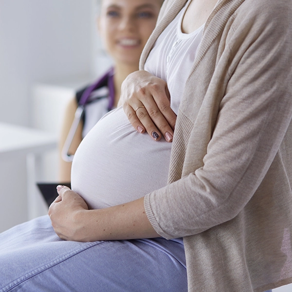 A pregnant woman gently cradles her rounded belly, seated in a medical office, with a healthcare provider in the background.